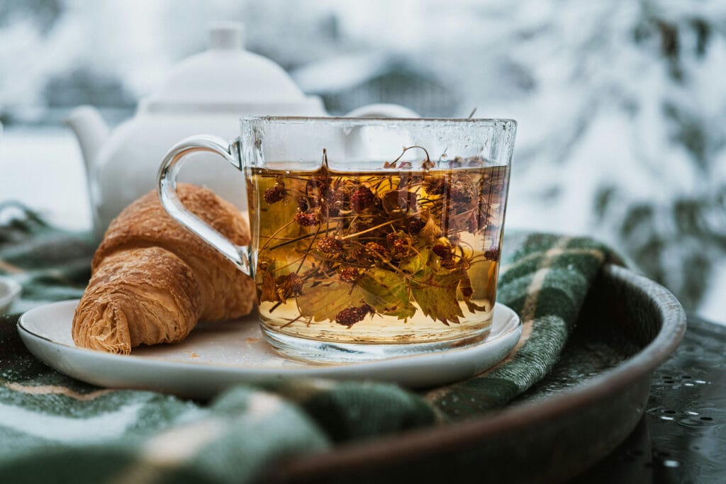 Herbata z suszonymi owocami w przezroczystym kubku na drewnianej tacy, obok croissant, na tle zimowego, śnieżnego krajobrazu, idealna na chłodne dni.