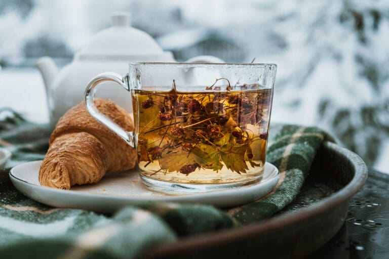 Herbata z suszonymi owocami w przezroczystym kubku na drewnianej tacy, obok croissant, na tle zimowego, śnieżnego krajobrazu, idealna na chłodne dni.