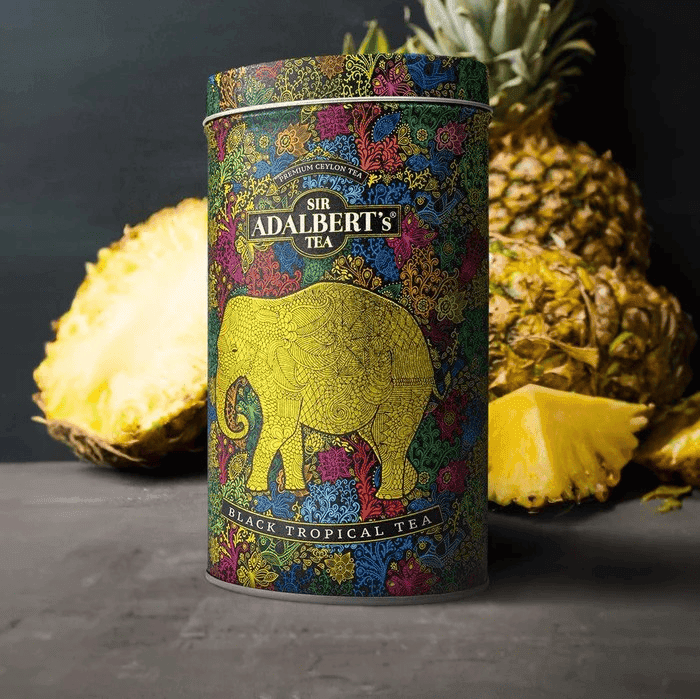 Adalberts Black Tea Tropical 110g Puszka 3 Aplikator ale nie widać, na zdjęciu popularyzowany produkt z czarnym, tropikalnym motywem i złotym słoniem, opakowanie herbaty Adalbert's Sir, otoczone świeżymi ananasami, dla miłośników luksusowej herbaty czarnej tropikalnej.