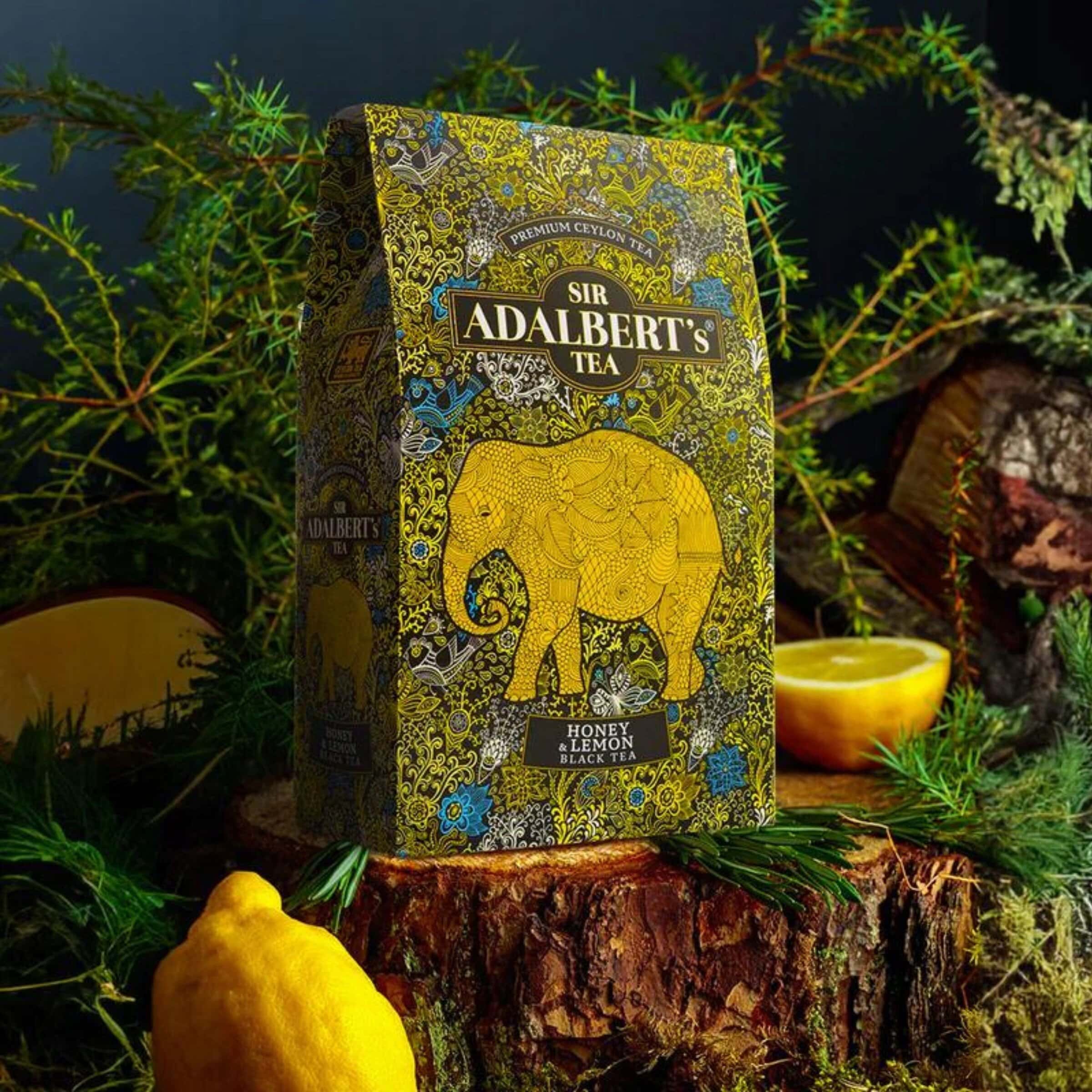 Herbata ADALBERT'S TEA BLACK TEA HONEY & LEMON 5 Wyraziste zdjęcie herbaty Sir Adalbert's Honey & Lemon Black Tea w ozdobnym opakowaniu z motywem słonia, ustawione na pniu drzewa w otoczeniu zieleni i cytrusów, idealne do prezentacji luksusowych herbat.