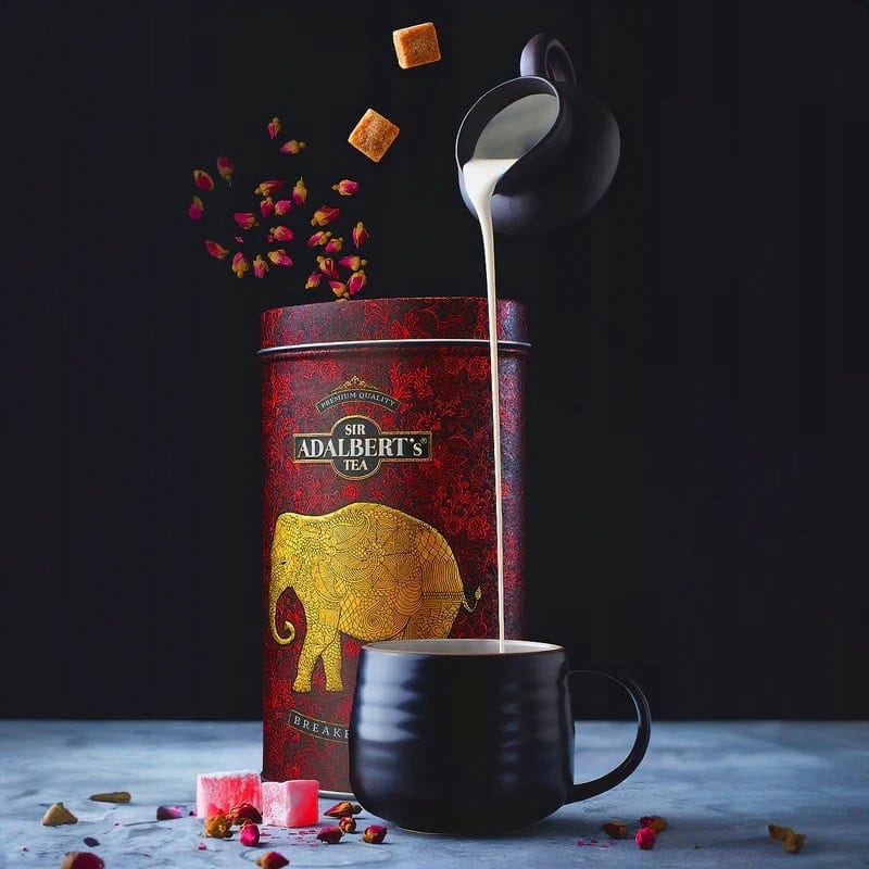Czarna filiżanka z mlekiem na stole, obok czerwone pudełko z herbatą Sir Adalbert's Tea i płatki róż, cukierki i przyprawy, elegancka prezentacja herbaty.